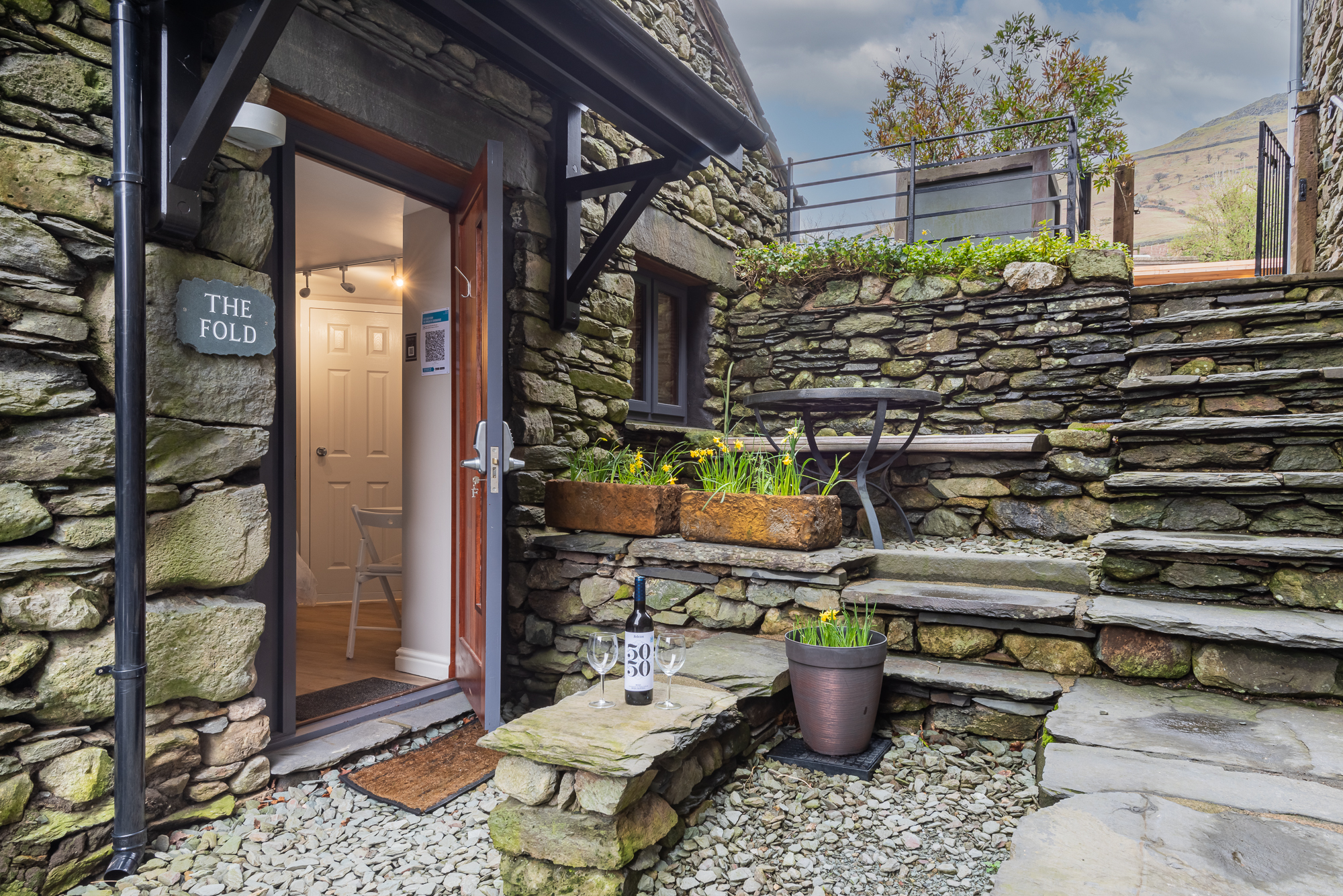Stone courtyard entrance to The Fold cottage at The Yan in Grasmere, featuring slate steps, flowers, and a bottle of wine on the patio.